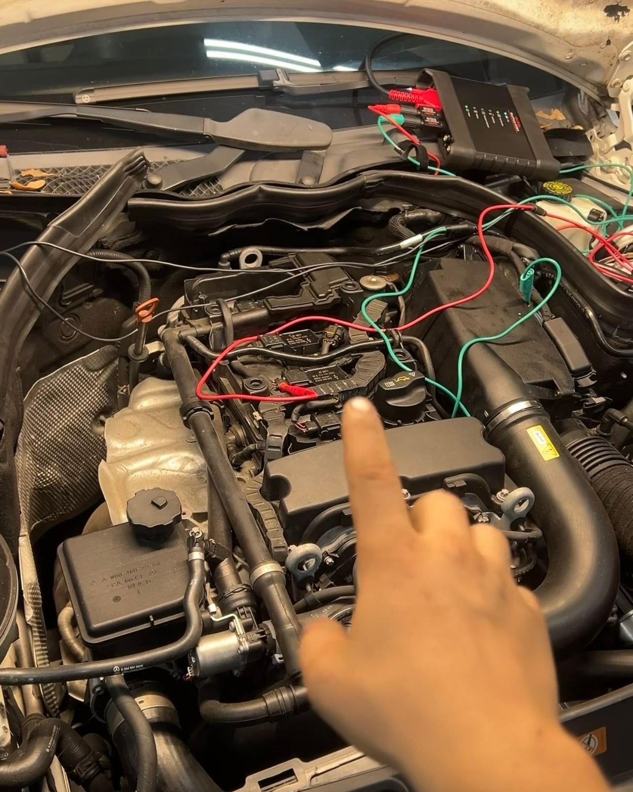 Oscilloscope test leads connected to engine wiring under the hood, with the scope module on the cowl and a technician pointing to the test point
